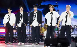 Five young men in black suits and white shirts with black ties perform on stage.