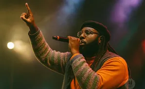 A Black man in a beret and glasses performs into a microphone, raising his hand in the air.