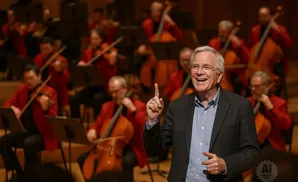 A man in a suit smiles and points while an orchestra plays behind him.