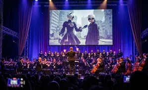 An orchestra and choir perform in front of a large screen displaying characters from the game NieR: Automata.