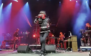 A band performs on stage with blue and red lights, a singer in a striped sweater in the foreground.