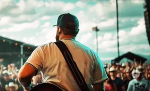 Man with guitar on stage, facing crowd with arms raised, under a cloudy sky.