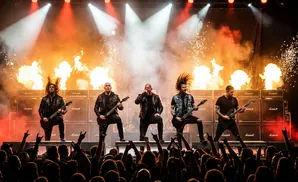 A metal band performs on a stage with fire and sparks erupting behind them, as an excited crowd cheers.