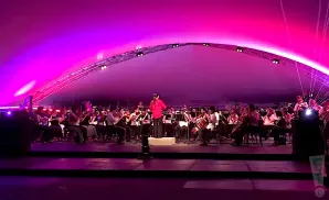 An orchestra performs under a large, pink, arched tent, illuminated by purple lights.