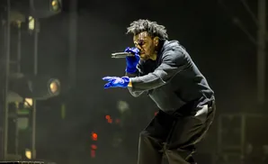 A singer in blue gloves holds a microphone and sings on a dark stage.