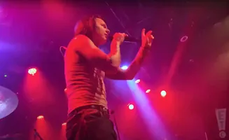 A male singer with long hair performs on a dimly lit stage, holding a microphone and gesturing with his hands, surrounded by red and purple stage lights.