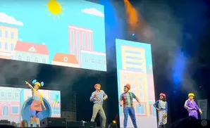 A live performance with costumed characters in front of a cityscape backdrop and bright lights.