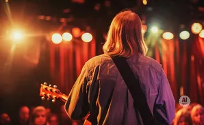 Guitarist on stage with red curtains and lights, playing for a blurred audience.