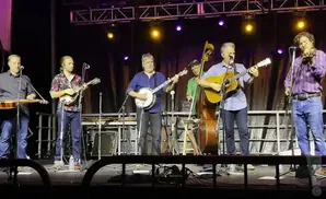 Band performs bluegrass music on stage with various instruments including guitar, banjo, mandolin, and bass.