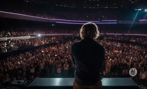 A lone performer with blonde hair stands on a stage, facing a massive, cheering audience in a dimly lit stadium.
