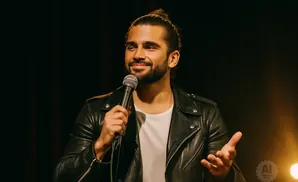 A man with dark hair in a bun, wearing a black leather jacket, smiles and holds a microphone.
