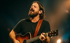A man with long brown hair and a beard plays an acoustic guitar on stage, eyes closed in song.