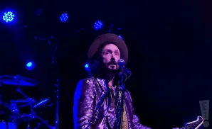 Man with hat and patterned jacket sings into microphone on a dark stage with blue lights.
