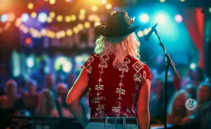 Woman in a cowboy hat and red patterned shirt on stage facing an audience.