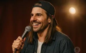 A man with long hair, a baseball cap, and a denim shirt laughs into a microphone.