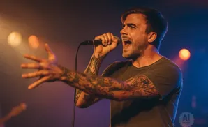 A tattooed man sings into a microphone with his arm outstretched on a dimly lit stage.