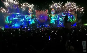 Fireworks erupt over a large crowd at a nighttime festival, with a fantastical stage featuring mermaid-like figures and a giant flower.