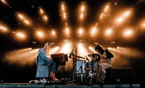 Two musicians perform on a stage lit by warm spotlights. A woman plays a keyboard on the left, and a person plays drums on the right.