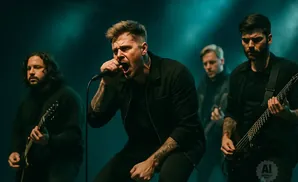 A male singer in black, with tattoos, sings into a microphone on stage with his band.
