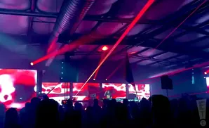 A crowd watches a DJ perform with red laser lights and a large screen displaying abstract red patterns.