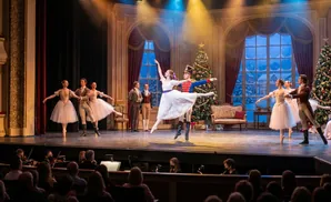 Dancers in ballet costumes perform on a stage with Christmas decorations, in front of an audience.