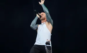 A bald man with tattooed arms sings into a microphone on stage, wearing a torn white tank top and black pants.