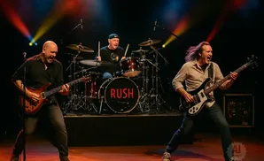 Rush band members perform live on stage. A guitarist, drummer, and bassist play their instruments.