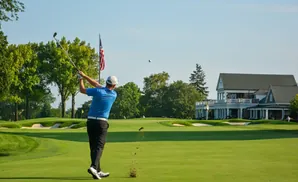 Golfer swings club on a sunny golf course with a clubhouse in the background.