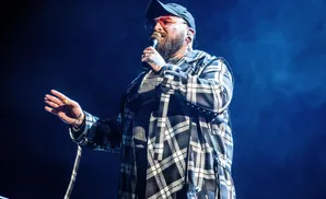 A man in a plaid jacket and cap sings into a microphone on a dark stage.