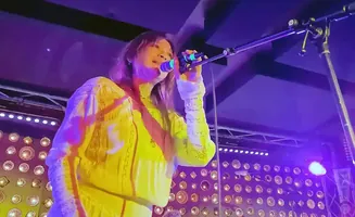 Singer in a yellow dress performs on stage, holding a microphone under purple lighting.