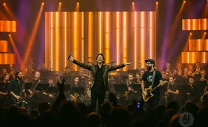 A conductor with arms outstretched, leads a band on stage with a backdrop of illuminated vertical panels.