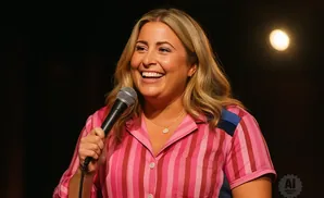A blonde woman in a pink striped shirt holds a microphone and laughs.