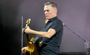 Bryan Adams on stage playing an acoustic guitar.