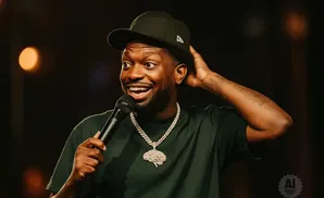 Comedian holding a microphone and touching his head while wearing a hat and a brain pendant.