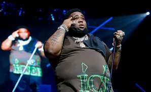 Rapper Rod Wave performs on stage wearing a brown t-shirt with green lettering, large diamond chains, and rings.