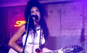 Woman with curly hair sings into a microphone while playing a guitar on a stage with neon lights.