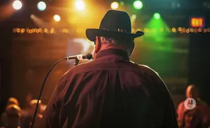 Man in a black hat on stage, facing away from the camera, speaking into a microphone, with audience and stage lights in the background.