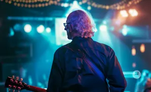 Musician with curly white hair and glasses, seen from behind, playing a guitar on a brightly lit stage.