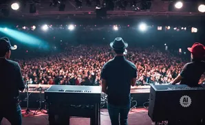 Three musicians on stage facing a large, excited audience under concert lighting.