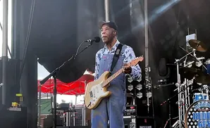 Buddy Guy performs on stage wearing a floral shirt and overalls, playing a light-colored electric guitar.