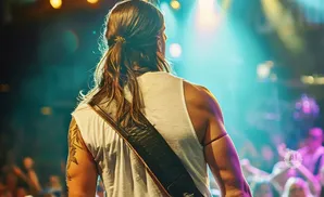 A man with long hair tied back, wearing a white tank top and guitar strap, stands on a stage with a crowd in the background.