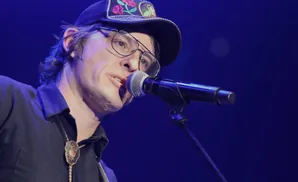 A man in a black shirt and hat sings into a microphone on a blue-lit stage.