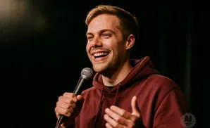 A man with blond hair laughs into a microphone, wearing a maroon hoodie.