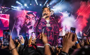 A rapper performs on stage with a large screen behind him, as an audience holds up phones to record.