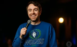 A man in a blue sweatshirt with a wolf logo smiles holding a microphone on a dark stage.