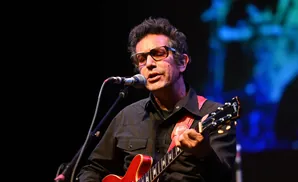 A man with glasses playing a red electric guitar and singing into a microphone on stage.
