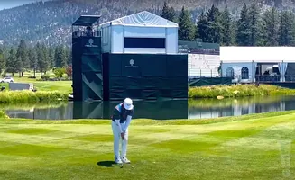 Golfer putting on a green with a large tent and scoreboard in the background.