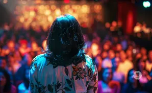 Person on stage with a floral shirt facing a blurry, cheering audience under colorful lights.