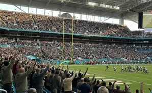 Fans cheer at a football game in a stadium, with players on the field and a large screen showing replays.