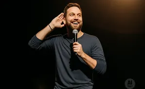 A man with a beard holds a microphone and cups his ear, smiling under a spotlight.